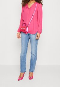 Blouse rose à manches longues, jeans bleu clair et escarpins roses à bout pointu avec des accents en forme de cœur ; petit sac rose avec un détail en nœud.
