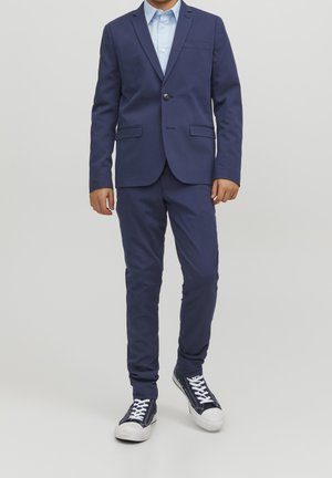 Marineblauer Anzug mit schlankem Schnitt, Ein-Knopf-Verschluss, zwei Vordertaschen, kombiniert mit einem hellblauen Hemd und legeren marineblauen Sneakern.