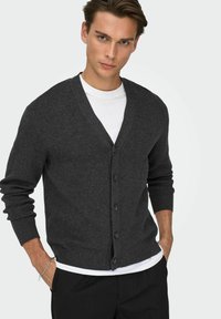 Cardigan grigio scuro in tessuto morbido, con scollo a V, chiusura con bottoni, maniche lunghe e taglio rilassato. Indossato sopra una camicia bianca.