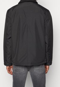 Sort letvægtsjakke med ribbet krave, lige snit og minimalt design, båret over grå jeans.