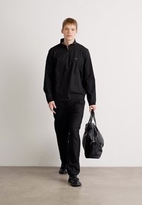 Čierna ľahká vetrovka na zips, kombinovaná s ladnými nohavicami a čiernymi topánkami. Nesie elegantnú čiernu športovú tašku. Jednoduchý, moderný dizajn.