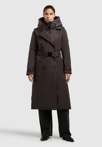 Donna che indossa un lungo trench coat marrone scuro con cintura e un interno imbottito con cappuccio, pantaloni neri e scarpe nere a punta, vista frontale.