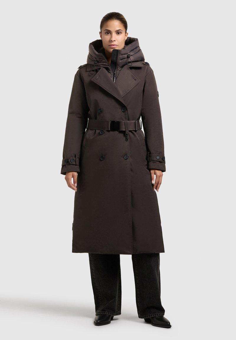 Donna che indossa un lungo trench coat marrone scuro con cintura e un interno imbottito con cappuccio, pantaloni neri e scarpe nere a punta, vista frontale.