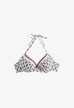 Frau mit langen dunklen Haaren, die einen gemusterten Bikini trägt, steht an einem sonnigen Strand mit großen Felsen im Hintergrund.