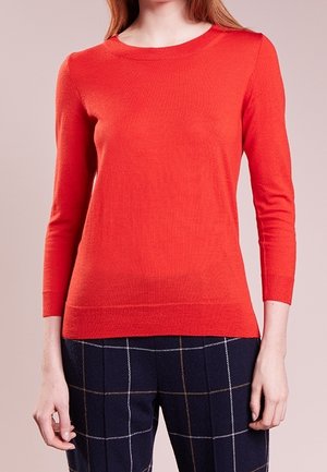 Kvinde iført en tætsiddende rød langærmet sweater og mørkeblå ternede bukser med hvide og brune linjer på en neutral baggrund.