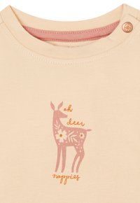 Beige Hemd mit einem rosa und weißen Rehdesign, das Blumen und den Text "oh deer nappies" zeigt. Enthält einen Druckknopf am Kragen.