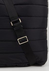 Sac en tissu noir matelassé avec sangle noire réglable et détail de boucle rectangulaire dorée.