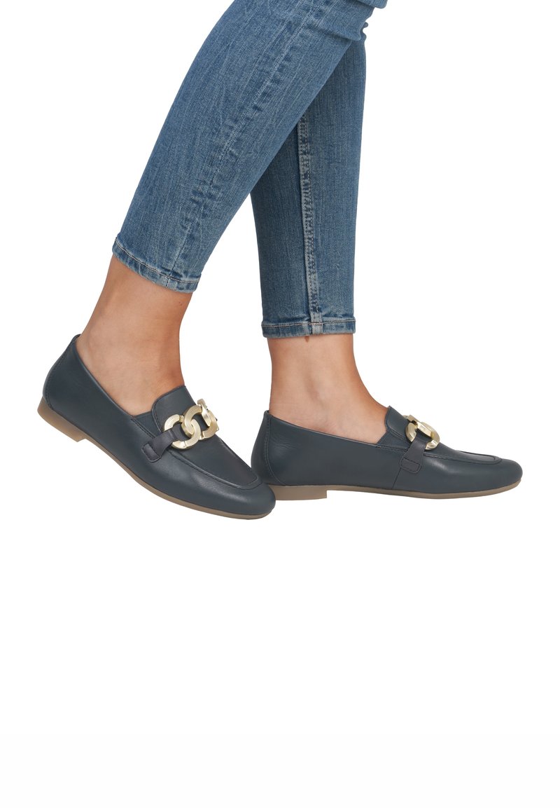Pieds portant des mocassins en cuir bleu foncé avec un détail chaîne dorée, associés à un jean skinny bleu retroussé.