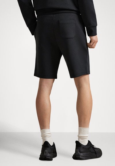 Black polyester shorts with a back pocket, featuring a relaxed fit and contrasting texture. Model wears black sneakers and white crew socks.