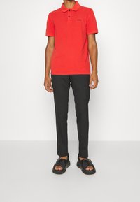 Man som bär en röd kortärmad polo skjorta med svart logotyp, svarta slim-fit byxor och svarta öppna sandaler mot en vit bakgrund.