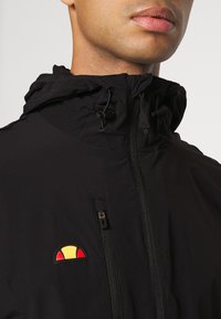 Chaqueta negra con capucha, cierre de cremallera y un logotipo colorido. Textura suave con un diseño funcional y cordones ajustables.