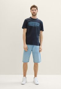 Mørkeblå t-shirt med rund hals og grafisk tryk i lysere blå; stylet med lyse denim shorts og hvide sneakers. Afslappet stil.