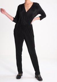 Svart plisserad jumpsuit med djup V-ringning, trekvart ärmar, samlad midja och avsmalnande ben. Slät textur utan synliga mönster.