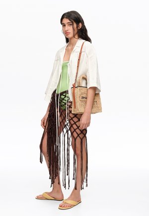 Mujer con chaqueta blanca corta sobre traje de baño verde, falda marrón de macramé, sandalias amarillas y sosteniendo un bolso tejido de paja con asa de cuero.