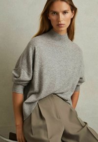 Jasnoszary sweter dziany z wysokim kołnierzem, luźnym krojem i ściągaczami na rękawach, noszony na beżowych, plisowanych spodniach. Gładka faktura, minimalne detale.