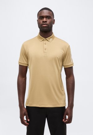 Mann trägt beiges Kurzarm-Polo mit Kragenlogo und schwarze Hose, steht vor einfachem weißem Hintergrund.