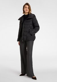 Giacca nera imbottita con cappuccio, chiusura a zip e tasche laterali, abbinata a pantaloni grigi a gamba larga e scarpe scure.