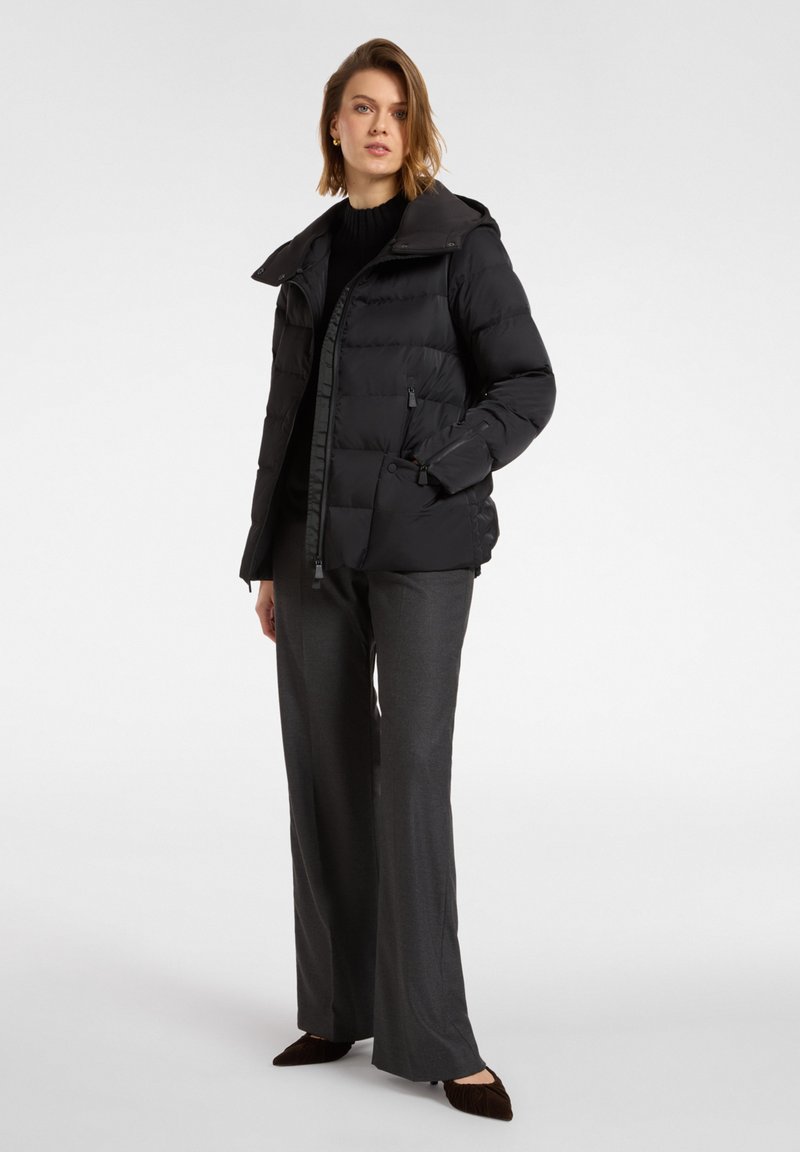 Giacca nera imbottita con cappuccio, chiusura a zip e tasche laterali, abbinata a pantaloni grigi a gamba larga e scarpe scure.