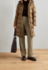 Persoon in een lange bruine pufferjas, olijfgroene broek, zwarte laarzen, en met een zwarte tote bag, staand op een houten vloer.