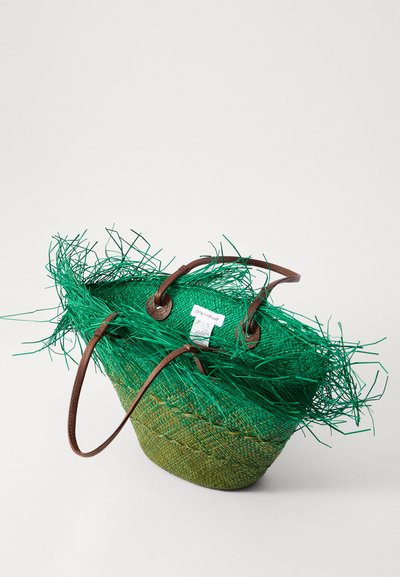 Grön vävd halm bucket bag med texturerad grön frans, bruna läderhandtag och en etikett fäst inuti.