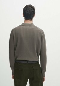 Brun ribbet sweater med krave, med lange ærmer og en tætsiddende pasform. Båret med mørkegrønne bukser, vist fra bagperspektiv.