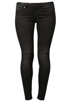 Femme marchant portant un haut gris à manches longues, un jean skinny noir, des bottes noires, portant un grand dossier ou un portfolio noir.