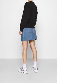 Persona con sudadera negra, falda de mezclilla azul con dobladillo deshilachado, calcetines blancos y zapatillas blancas de pie contra un fondo liso.