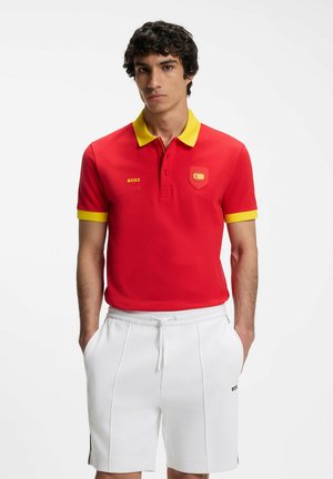 Młody mężczyzna w czerwonej koszulce polo z żółtym kołnierzykiem i mankietami, białe spodenki ze ściągaczem, stoi z rękami w kieszeniach.