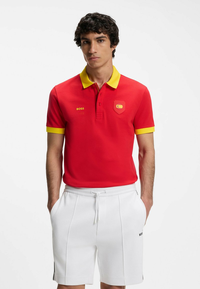 Jeune homme portant un polo rouge avec col et poignets jaunes, short blanc à cordon, debout les mains dans les poches.