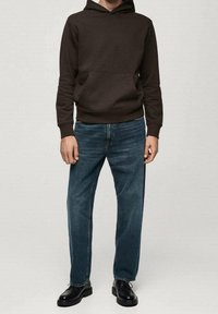 Uomo che indossa un hoodie marrone scuro, jeans dritti blu e scarpe di pelle nere, in piedi contro uno sfondo neutro.