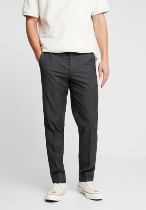 Mand iført mørkegrå slim fit bukser, hvide sneakers og en kortærmet off-white skjorte, stående med den ene hånd i lommen.