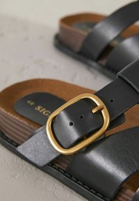 Zwarte leren sandalen hebben twee brede banden, een bruine gestructureerde voetbed en een gouden rechthoekige gesp voor een verstelbare pasvorm.
