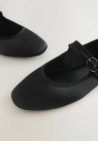 Ballerines noires en tissu lisse avec un bout arrondi et un bord élastique. Présentent une sangle avec une boucle circulaire en argent.