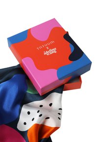 Scatola quadrata colorata con motivi astratti rosa, blu e rossi che contiene una sciarpa di seta con motivo blu e arancione con accenti neri.