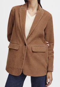 Brun kordfløyelsblazer med snittade slag, två framfickor och en enda knappknäppning, matchad med en krämfärgad V-ringad tröja.