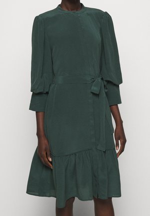 Abito verde scuro con maniche a sbuffo tre quarti, scollo rotondo, cintura in vita e orlo arricciato su una persona dalla pelle scura.