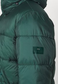 Grön puffjacka med glänsande yta, med kviltade sektioner, en logotyp som läser "PS" och "Paul Smith", samt en dragsko på huven.