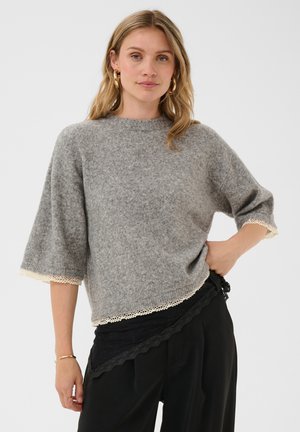 Femme debout avec une main sur la hanche, portant un pull gris à bordure en dentelle et un pantalon noir, sur un fond clair uni.