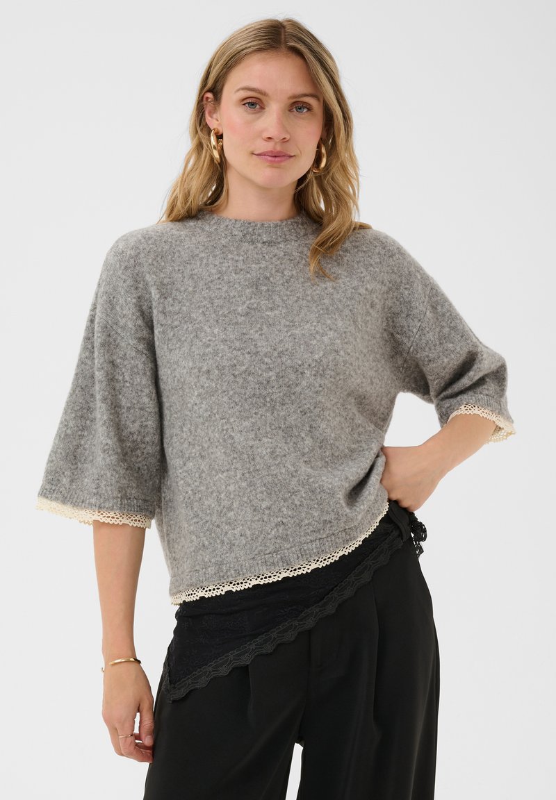 Femme debout avec une main sur la hanche, portant un pull gris à bordure en dentelle et un pantalon noir, sur un fond clair uni.