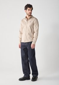 Camisa beige de botones con cuello, combinada con jeans azul oscuro. La camisa presenta un pequeño acento bordado. Zapatos negros completan el conjunto.