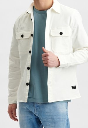 Mężczyzna w białej kurtce zapinanej na guziki, pod którą ma turkusową koszulę i jasnoniebieskie dżinsy, stoi na jednolitym tle.