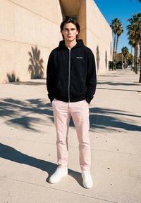 Ung mann står med hendene i lommene, iført en svart hettegenser, lyserosa bukser og hvite sneakers i en solrik bygate omgitt av palmer.