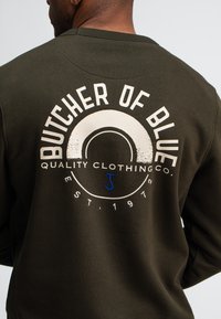 Donkergroene sweatshirt met een grafisch ontwerp op de achterkant, inclusief de tekst "BUTCHER OF BLUE" en een rond logo met accenten in crème en blauw.