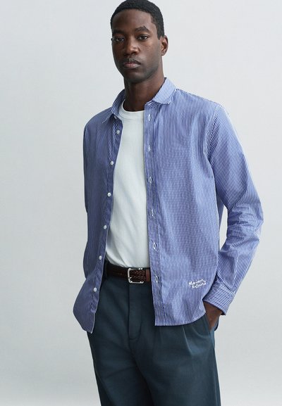 Camicia a righe blu e bianche con bottoni, tessuto leggero, calzata rilassata, maniche lunghe, abbinata a una t-shirt bianca e pantaloni scuri.