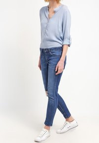Blusa azzurra a pois con colletto, maniche arrotolate, abbinata a jeans slim-fit blu e sneakers bianche.