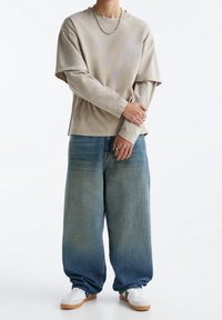 Personne portant des hauts à manches longues beiges superposés, un jean bleu ample, des baskets blanches, une chaîne argentée et des bagues aux deux mains.