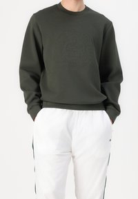 Sweat-shirt vert foncé avec un logo circulaire texturé sur le devant, associé à un pantalon blanc. Le tissu semble doux et décontracté.