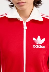 Rode zip-up jacket met een witte kraag en strepen. Heeft een opvallend wit adidas-logo op de borst in een sportief ontwerp.