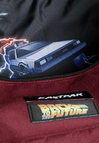 L'interno dello zaino presenta un tessuto nero con una grafica di una macchina DeLorean e motivi di fulmine, completato da un esterno color bordeaux e un'etichetta con il logo.