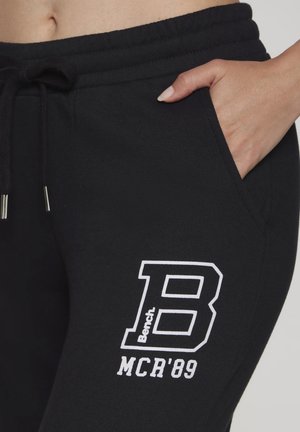 Nahaufnahme von schwarzen Jogginghosen mit Hand in der Tasche, Kordelzugbund und weißem "B Bench MCR'89" Logo am Oberschenkel.